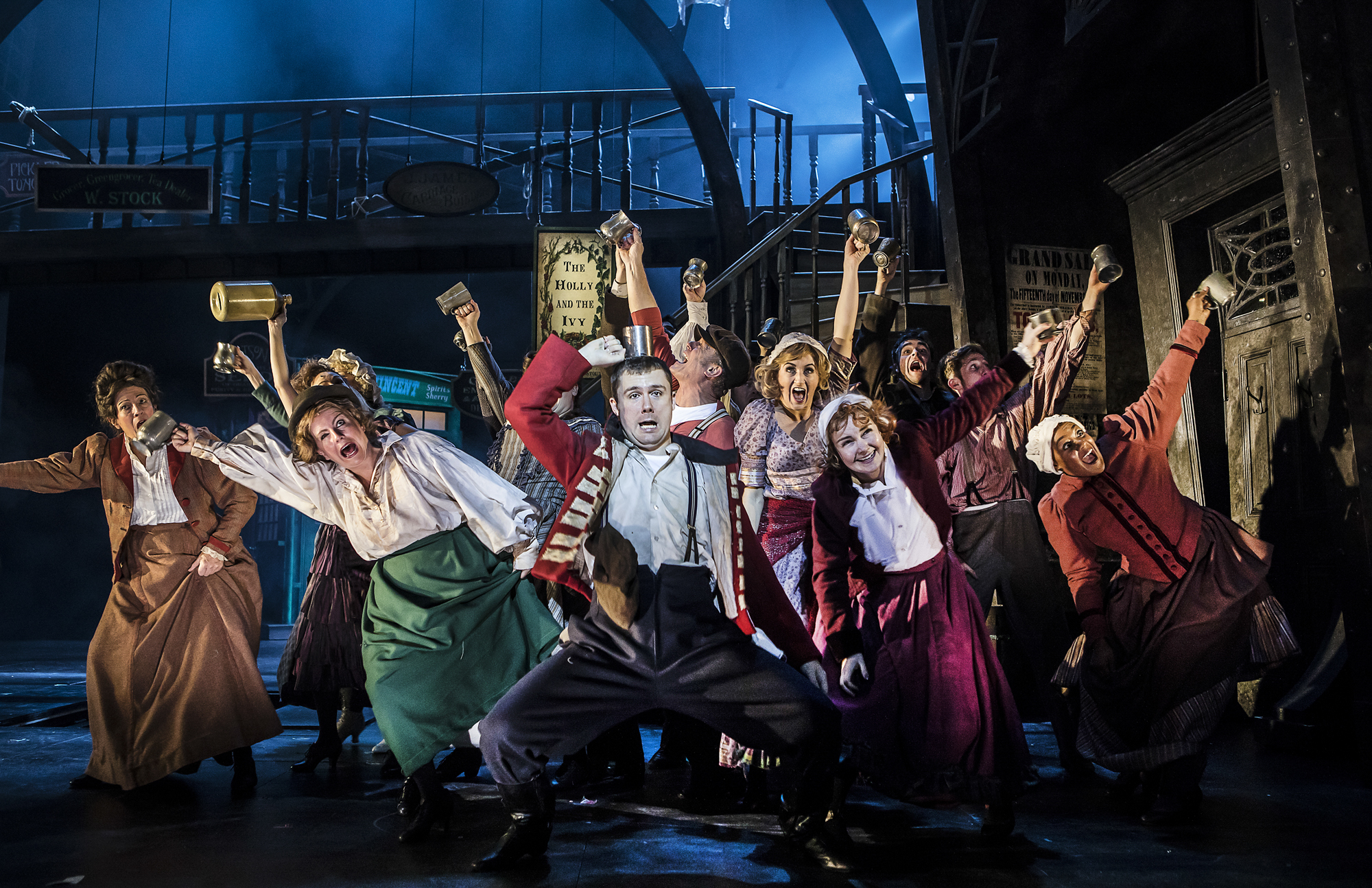 Production image from Scrooge. The company exclaim to the audience during a raucous pub scene on a darkly lit stage