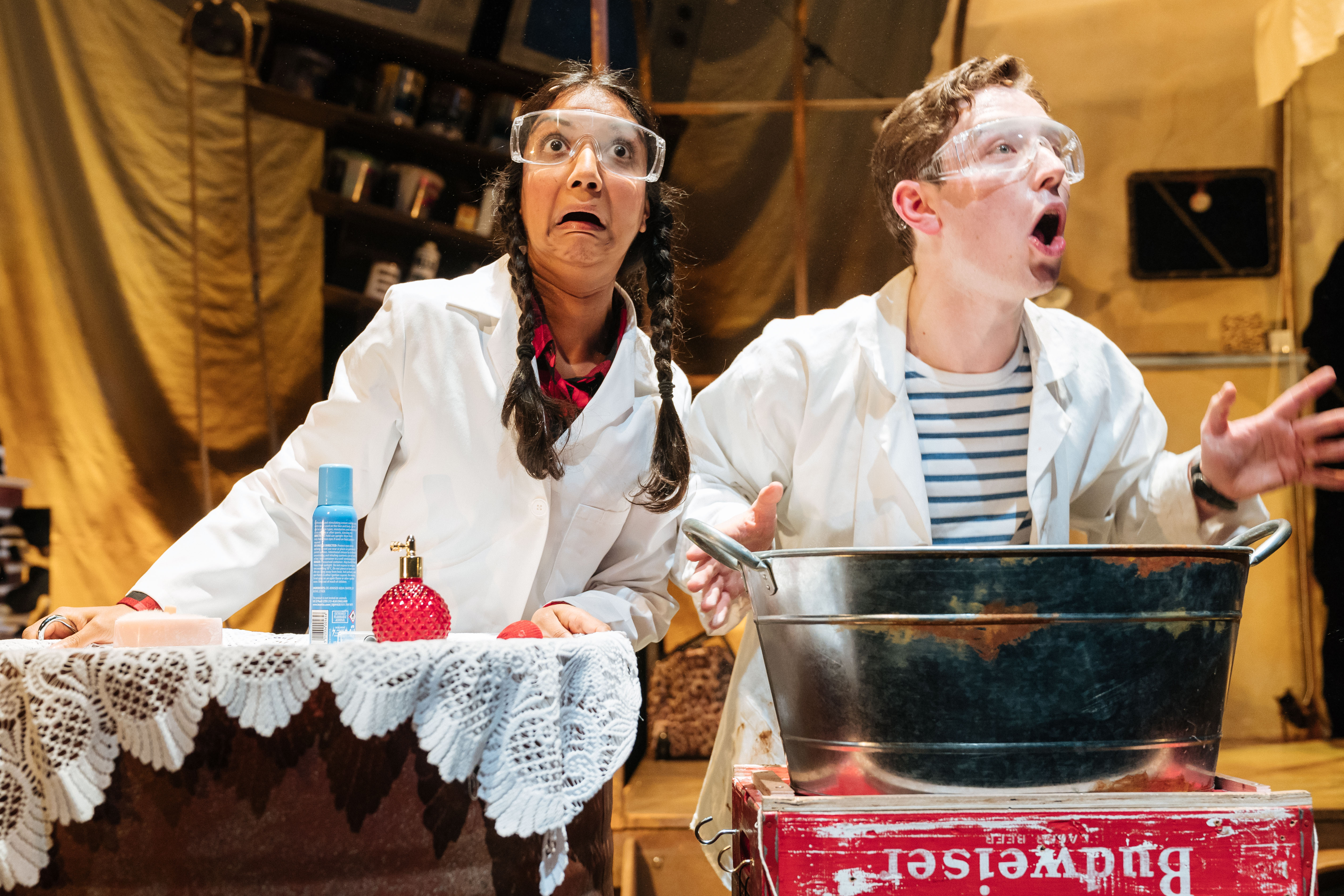 Production image from George's Marvellous Medicine. A scientist (Chandni Mistry) and George (Preston Nyman) exclaim to the audience as they mix a potion in a large steel basin, wearing white lab coats and clear safety goggles.
