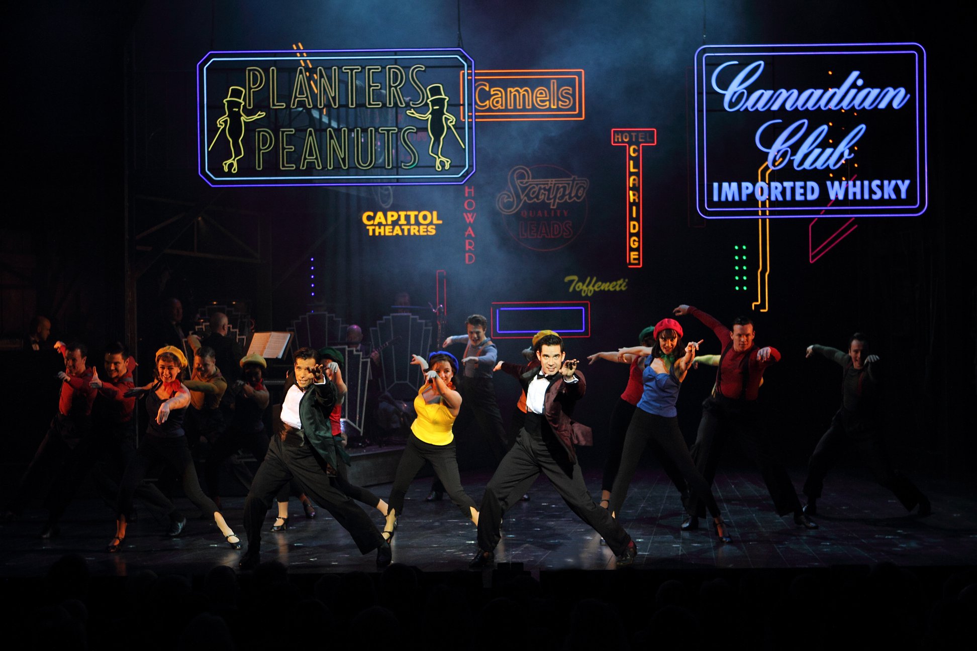 Production image from White Christmas. Danny Mac as Bob and Dan Burton as Phil lead the company in a dance number on stage, which is illuminated with neon signs. The pair are wearing suits.