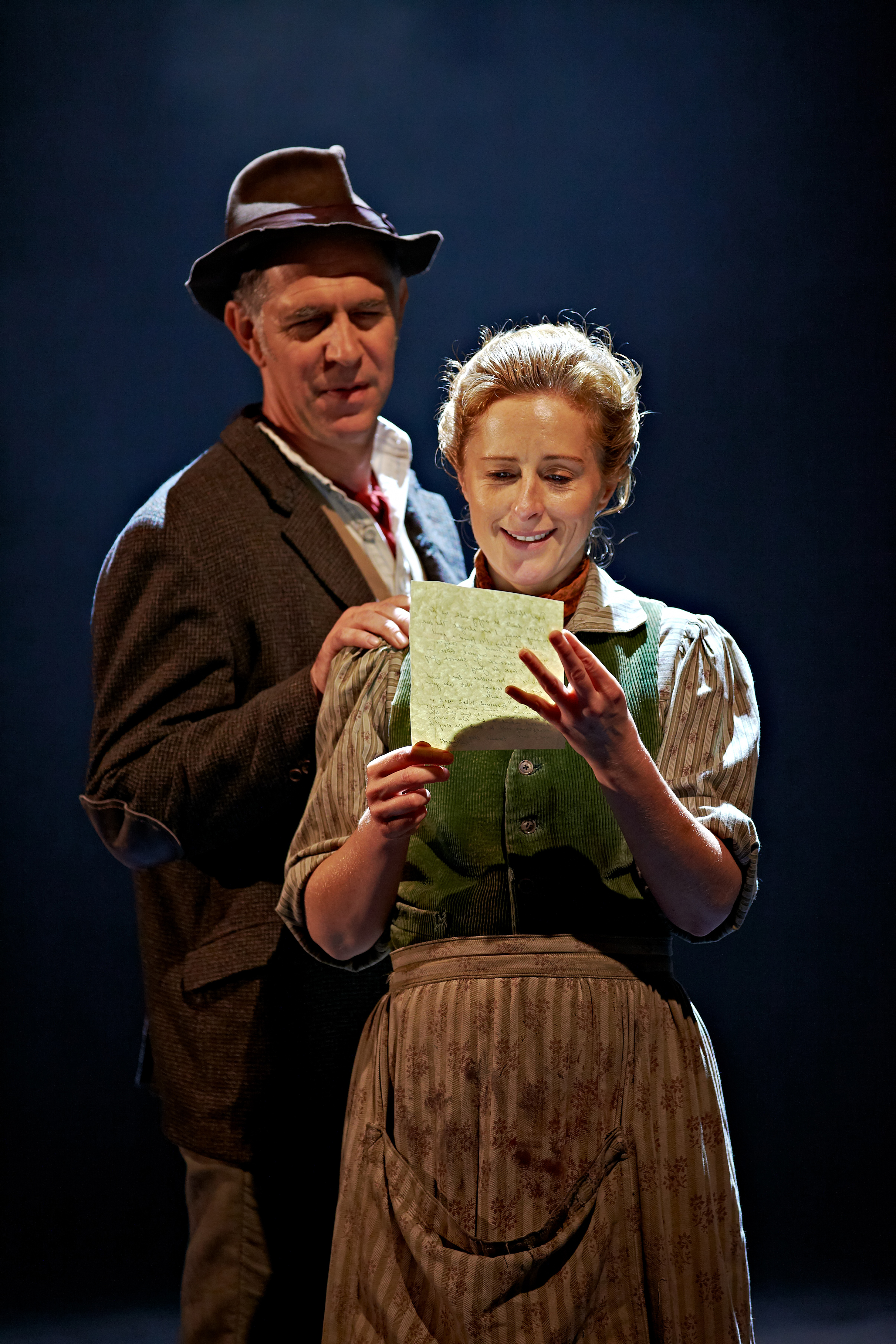 woman smiling whilst looking at the letter in her hand whilst a make standing close behind her comfortingly rests his hand on her shoulder, also looking at the letter and smiling.