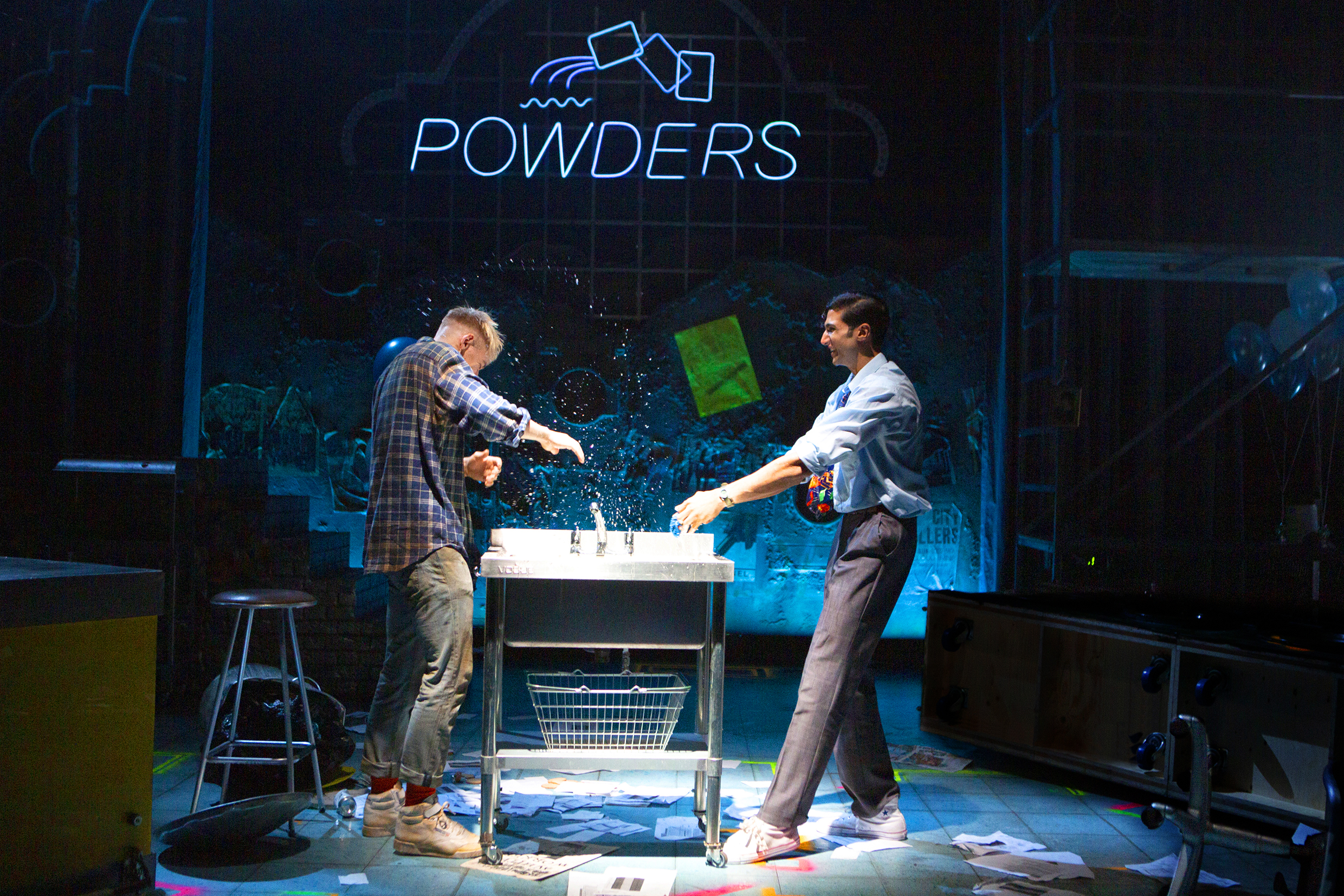 Production image from My Beautiful Laundrette. Johnny Fines as Jonny and Omar Malik as Omar splash each other with water from a drink on a darkly lit stage. A neon Powders sign is in the background.