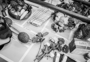 A black and white image of a table, upon which there is a bowl of fruit, a xylophone, maracas, a bunch of flowers, a flower garland and a box with the word award written on it. These are props and instruments used in the Giraffes Can't Dance rehearsal room.