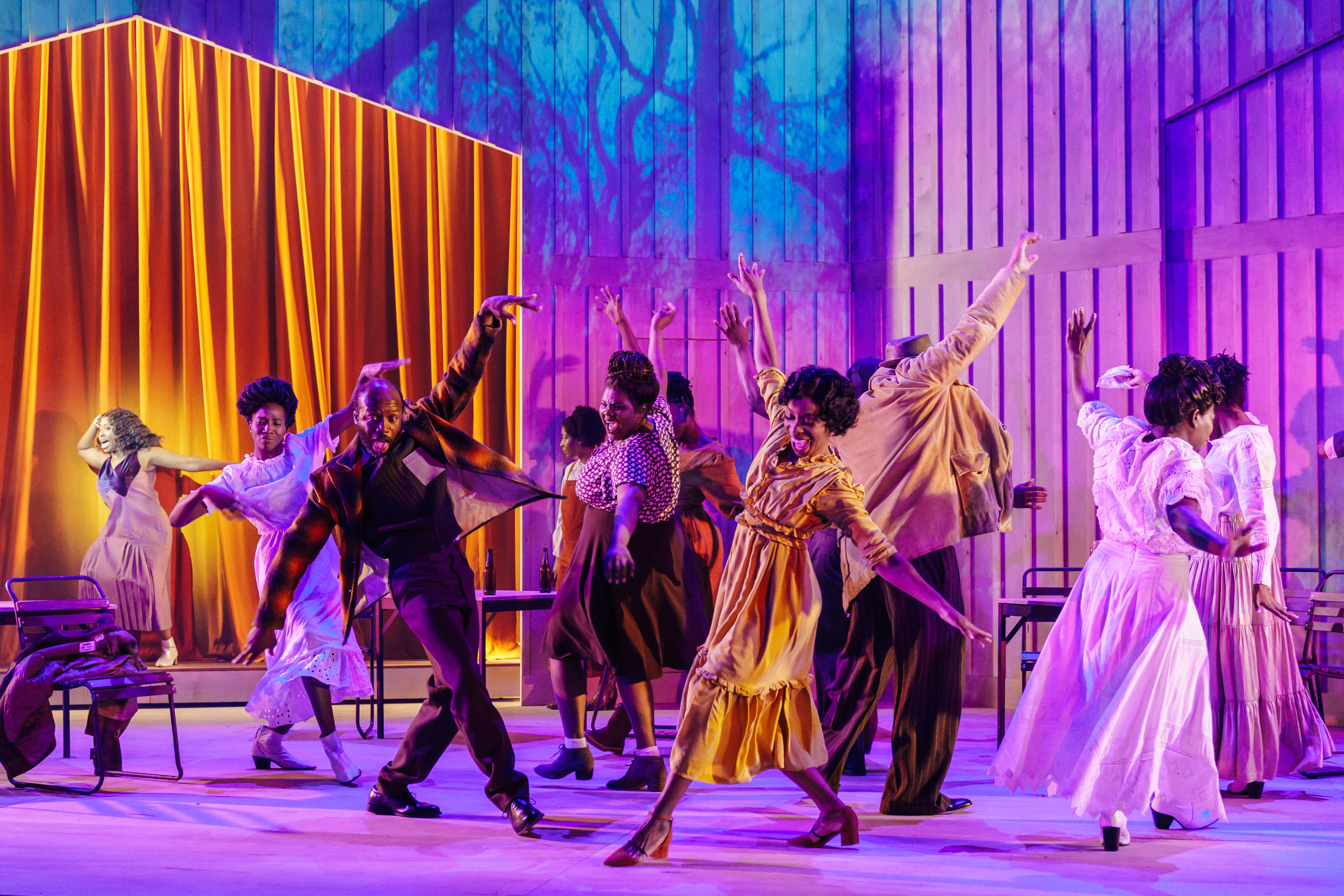 Production image from The Color Purple. The company dance in pairs on a purple and blue toned stage, with a golden curtain in the background.
