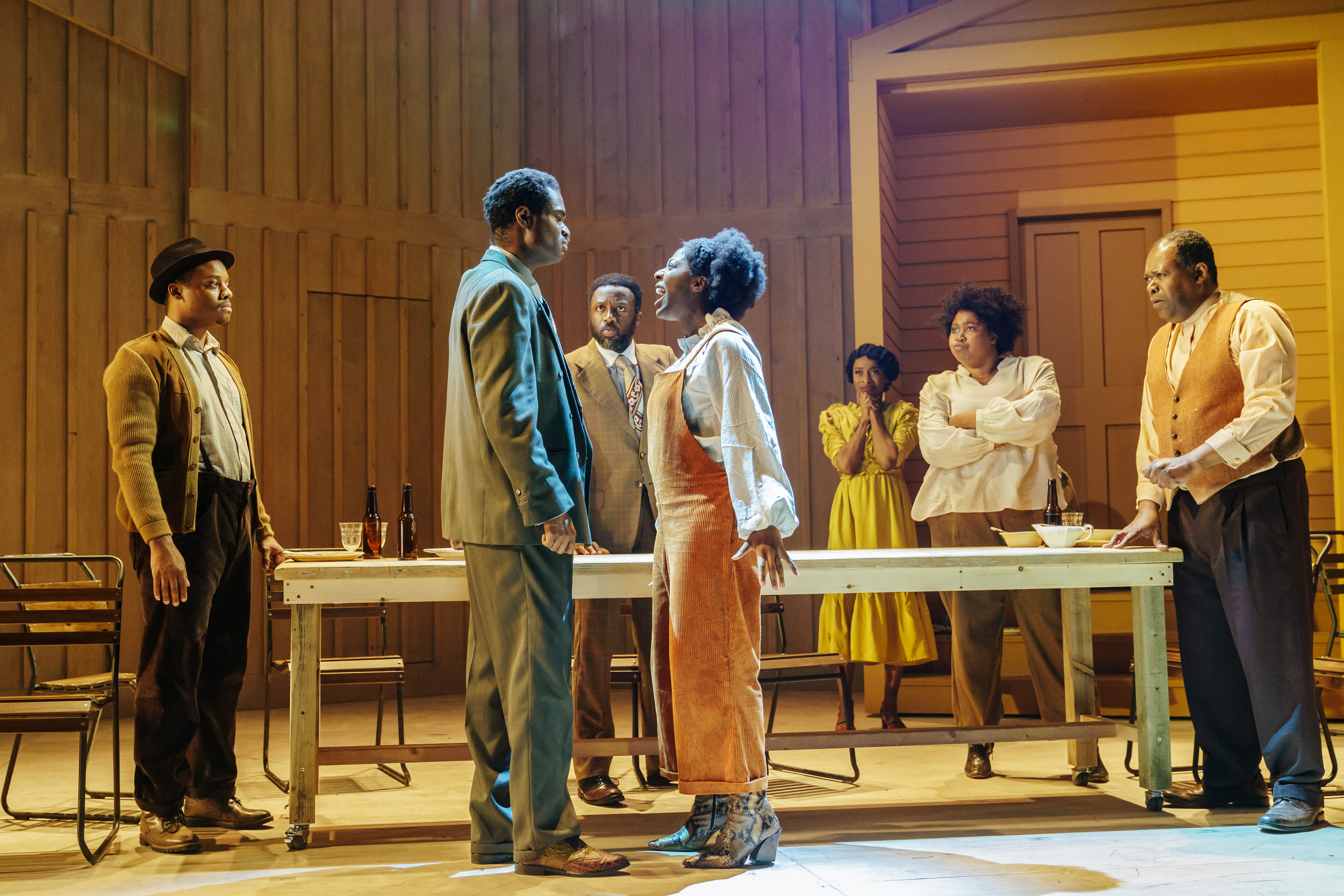 Production image from The Color Purple. Celie (T'Shan Williams) yells at Mister (Ako Mitchell) as five others watch on behind a table. The stage is lit in brown and gold hues.