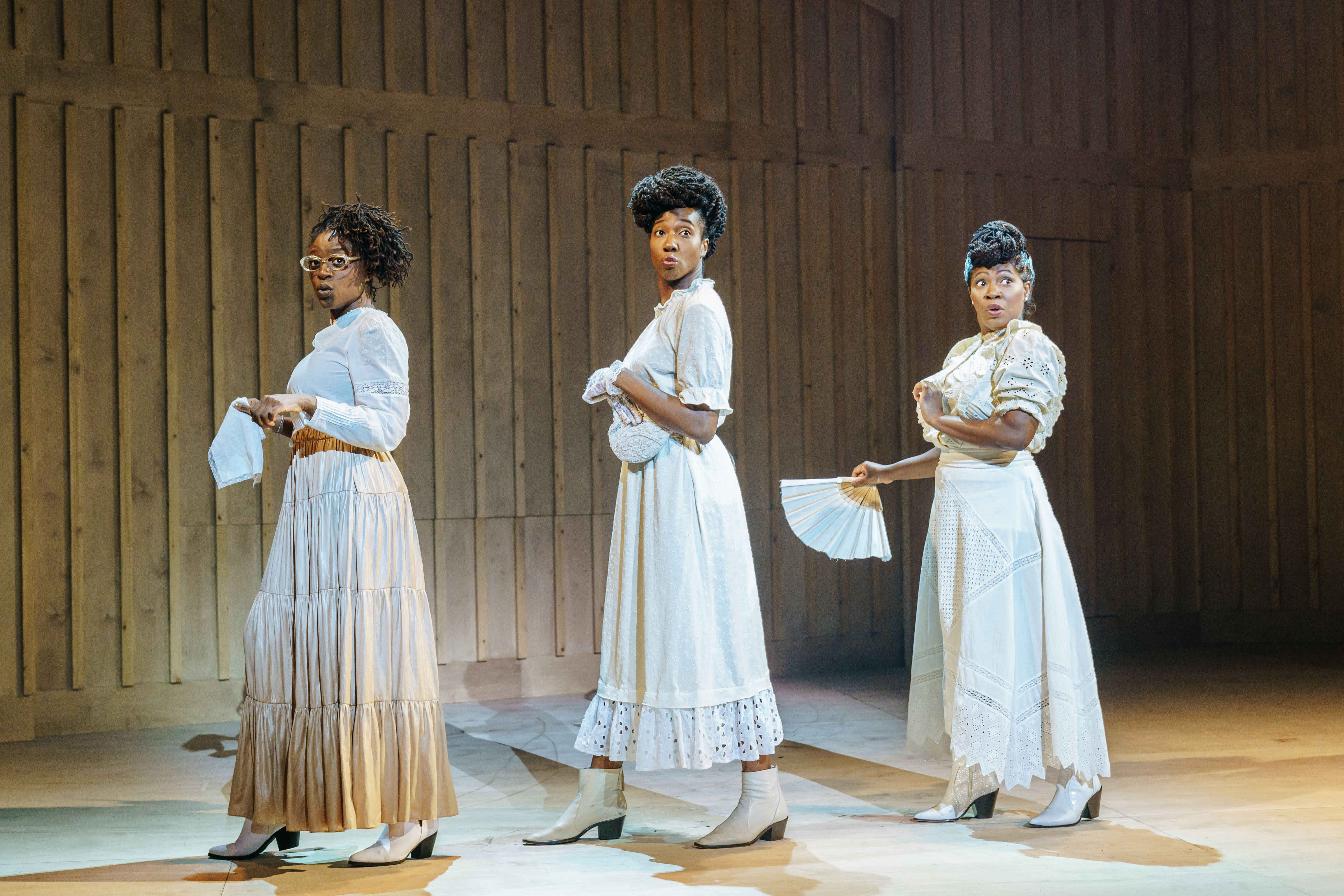 Production image from The Color Purple. Rosemary Annabella Nkrumah as Darlene, Danielle Kassaraté as Doris and Landi Oshinowo as Jarene stand on their sides singing in white, frilly cotton dresses against light panelling.