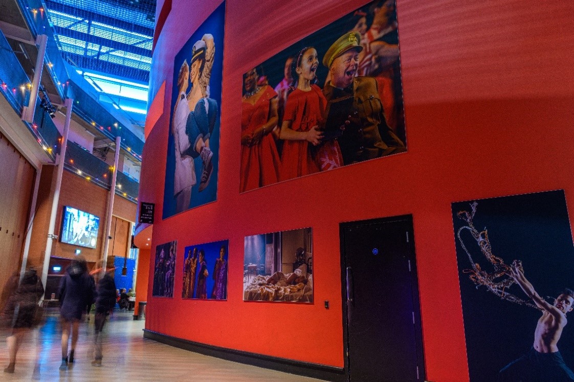 Part of Curve's public foyer. A large red wall with a black skirting board and doors is adorned with production pictures. here is a large screen in the distance and audience members are walking within the image.