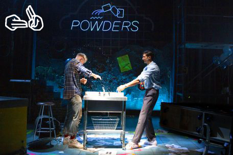 Production image from My Beautiful Laundrette. Johnny Fines as Jonny and Omar Malik as Omar splash each other with water from a drink on a darkly lit stage. A neon Powders sign is in the background.