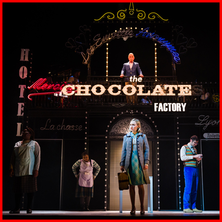 Production image from Romantics Anonymous. Angelique stands centre-stage looking into the distance, wearing a coat and carrying a suitcase. She is surrounded by three other characters including Jean-Rene, and overlooked from a balcony by another male. The dark stage is adorned in vintage, illuminated signage.