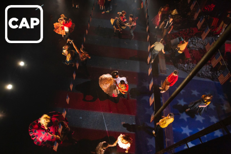Production image from West Side Story. The company dance in couples on a darkly lit stage adorned with USA flag bunting. The image is taken from above.