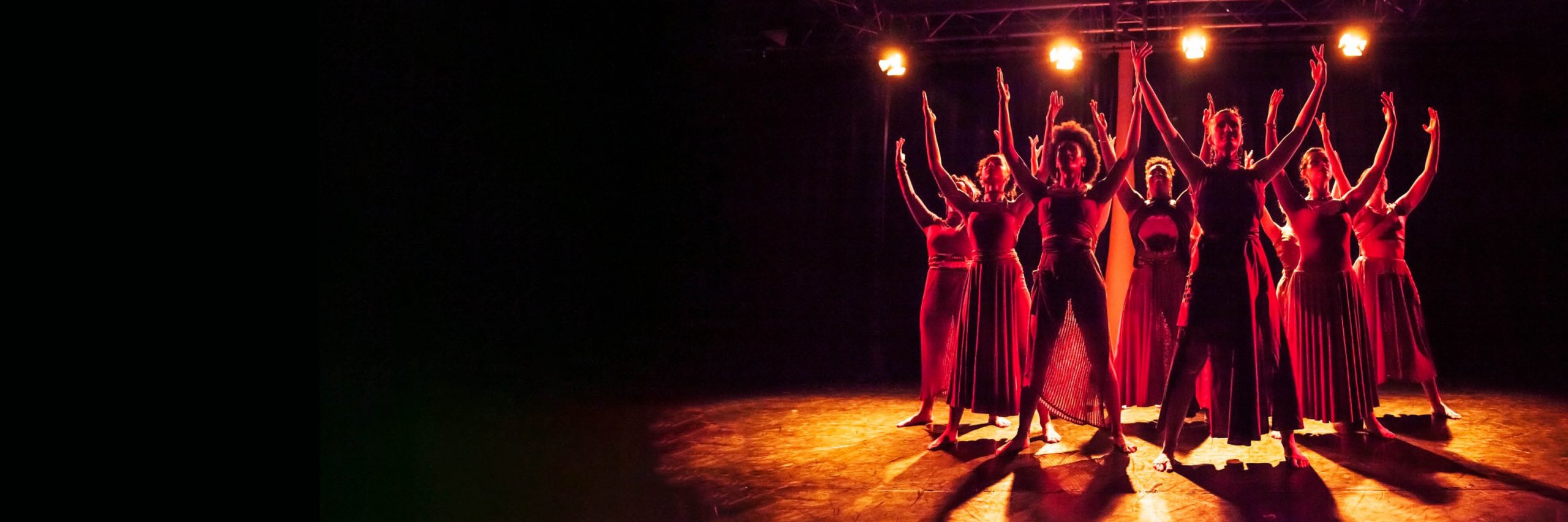 Promotional artwork for Spirit of Jazz: BOP Jazz Theatre Company. Seven dancers in various red outfits stand in a triangle formation with their arms in the air. They are on a stage, under golden spotlights.