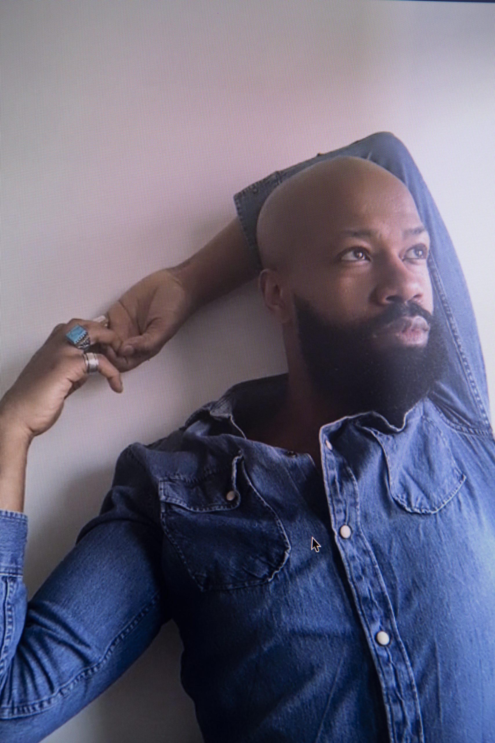 Gerrard Martin is a black man with a full beard. He is visible from the chest up and is wearing a denim blue shirt. He looks into the top right of the screen, beyond the image's frame and his arms are wrapped behind his head with his hands clasped together to the left.