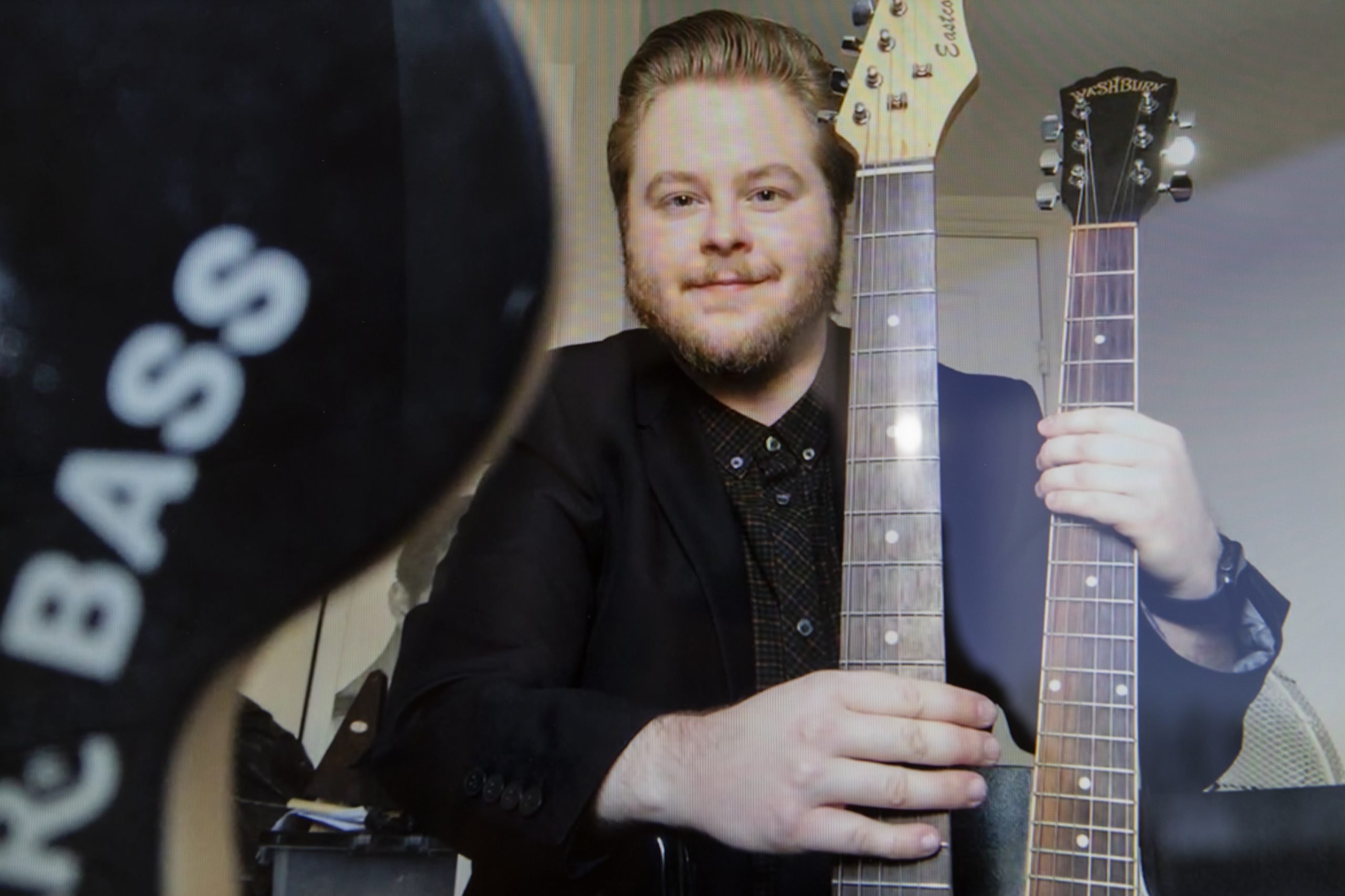 Jude Taylor is a white man with sandy blonde hair swept back off his face and a beard. He wears a black jacket and brown check shirt. To his right, he holds two guitars, their necks visible in the shot. To his left, a black object is at the forefront of the screen, with the word 'Bass' written on it.