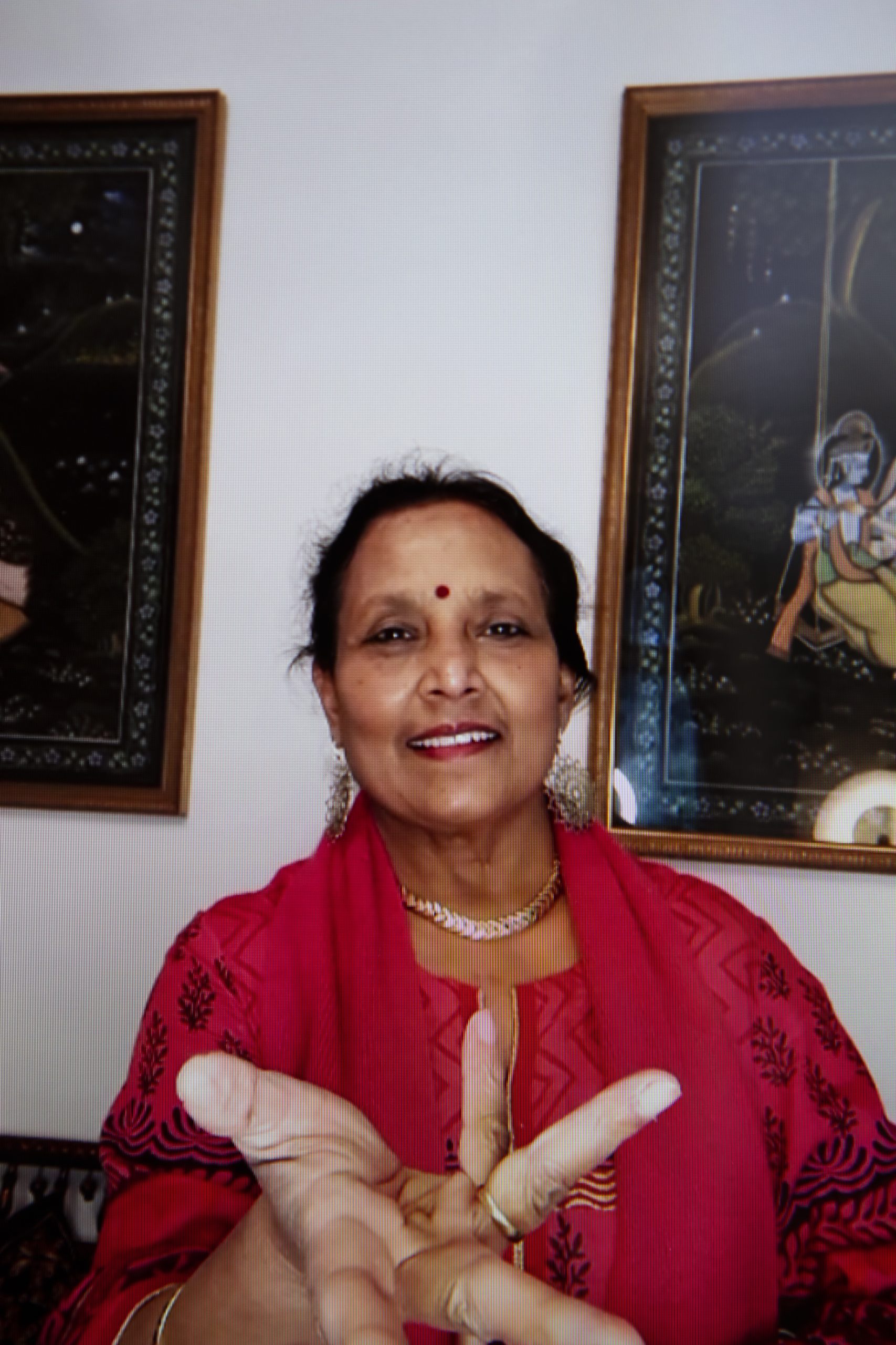 Nilima Devi smiles as she looks into the camera, her hand outstretched to the screen and fingers stretched to form a wheel shape. She wears a gold ring and necklace and her dark brown hair is tied away from her face. Nilima wears a red outfit which is decorated with gold and black embroidered patterns.