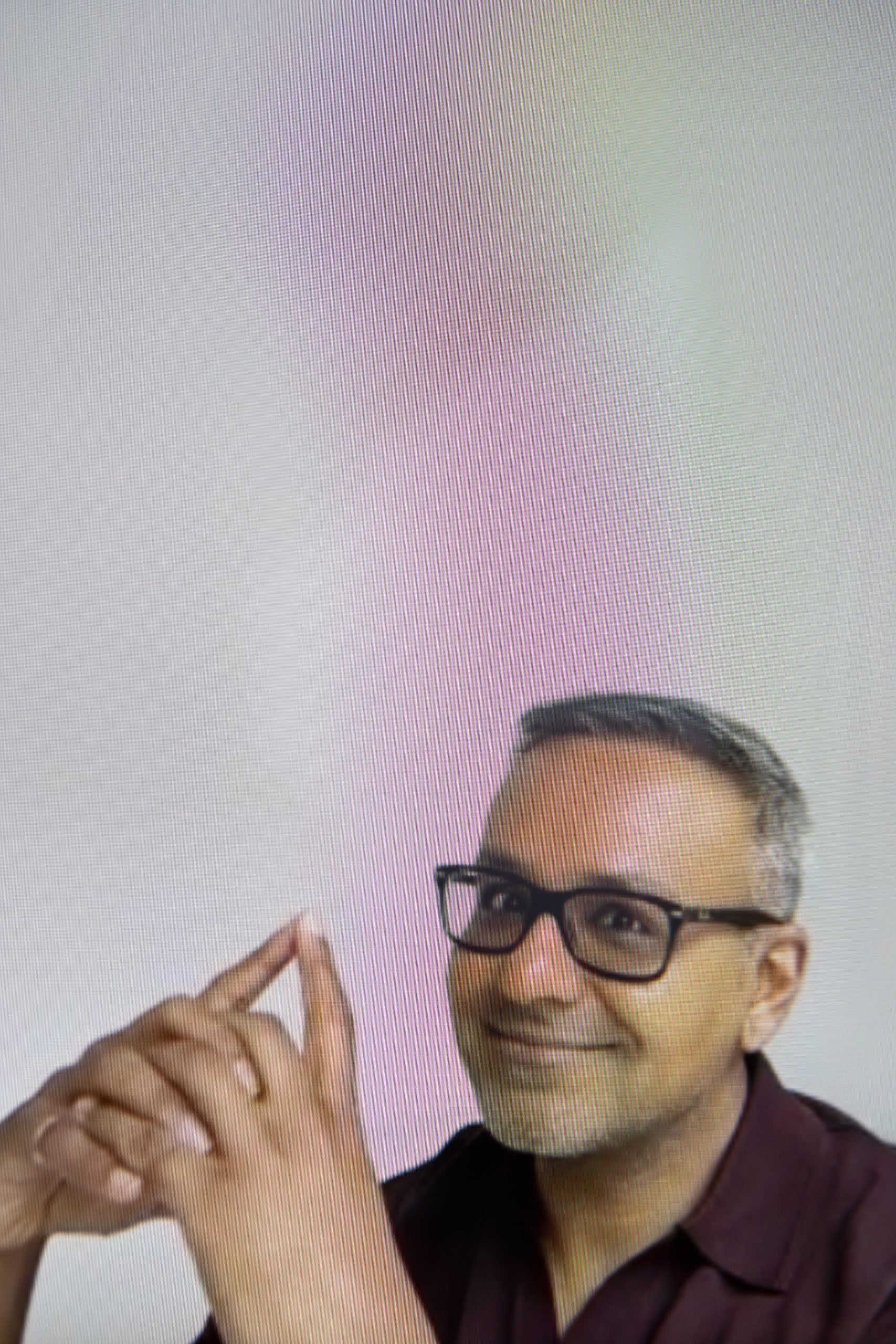 Samir Bhamra is visible from the chest up, in the bottom third of the portrait image. He wears a burgundy shirt and black glasses. He is an asian man with short grey hair and smiles as he looks into the camera. His arms are in front of his face, to the left and his hands are together, with his index fingers touching to make a triangle.