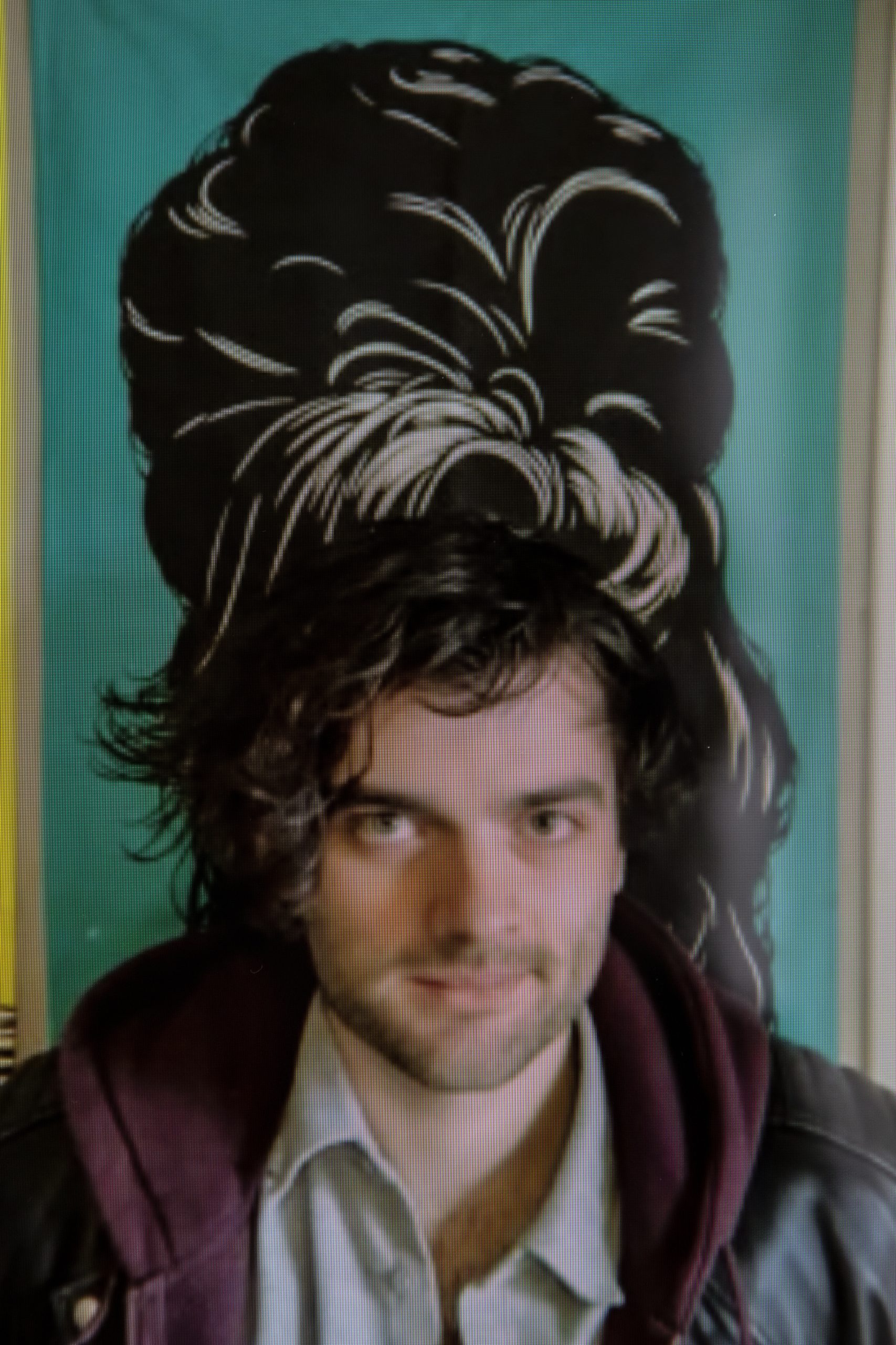 Samson Hawkins, a white man wearing a pale shirt and burgundy hoody with medium length brown hair swept across his face sits in front of a picture of Amy Winehouse's hair, creating the effect that it is an extension of his own.