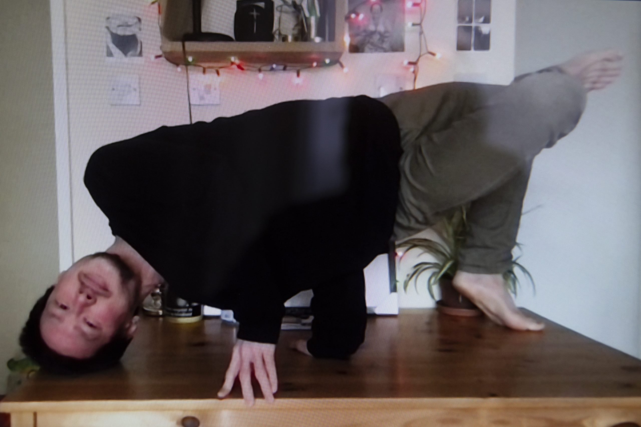 Si Rawlinson's full body is visible as he twists horizontally to balance on a table, supporting himself with his hands and one foot, with his other leg stretched upwards. His head is tipped upside down and rests on the table, he is looking into the camera with a calm expression.