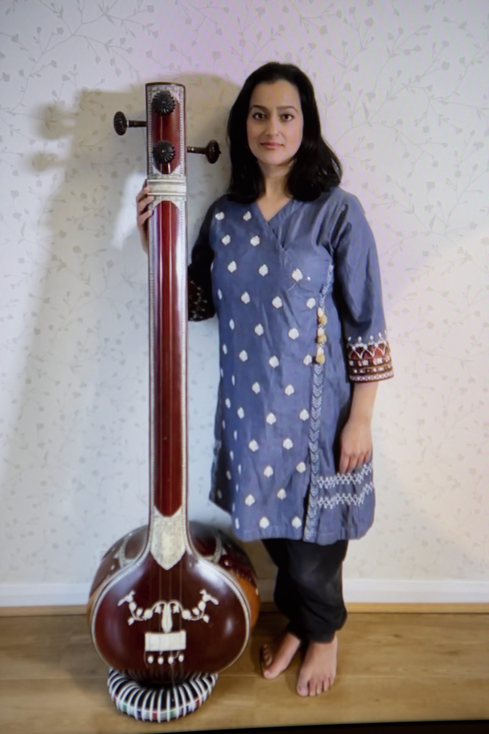 Sonia stands in the middle of the image, her full body visible. She is barefoot and wears a cornflower blue kurti with white embroidery on the front, she also wears black trousers which are visible from her knee to her ankle. Sonia is looking into the camera and smiling and her face is framed by dark shoulder length hair. To her left, she holds an upright sitar.