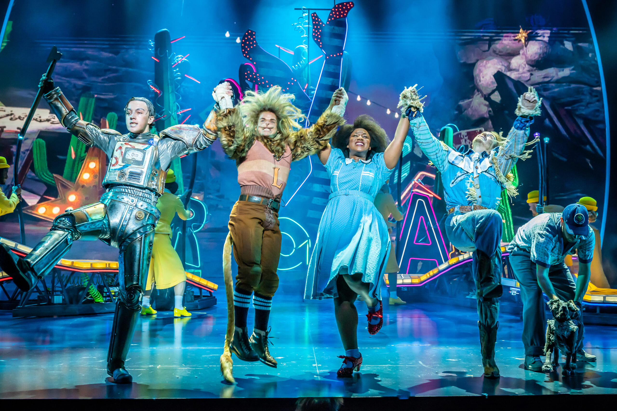 Production photograph from The Wizard of Oz. Tin Man (Paul French), Lion (Giovanni Spanó), Dorothy (Georgina Onuorah), Scarecrow (Jonny Fines) and Toto (Ben Thompson) leap into the air joyfully in front of neon signs on a blue stage. There is a white RP - Relaxed Performance - logo top left).