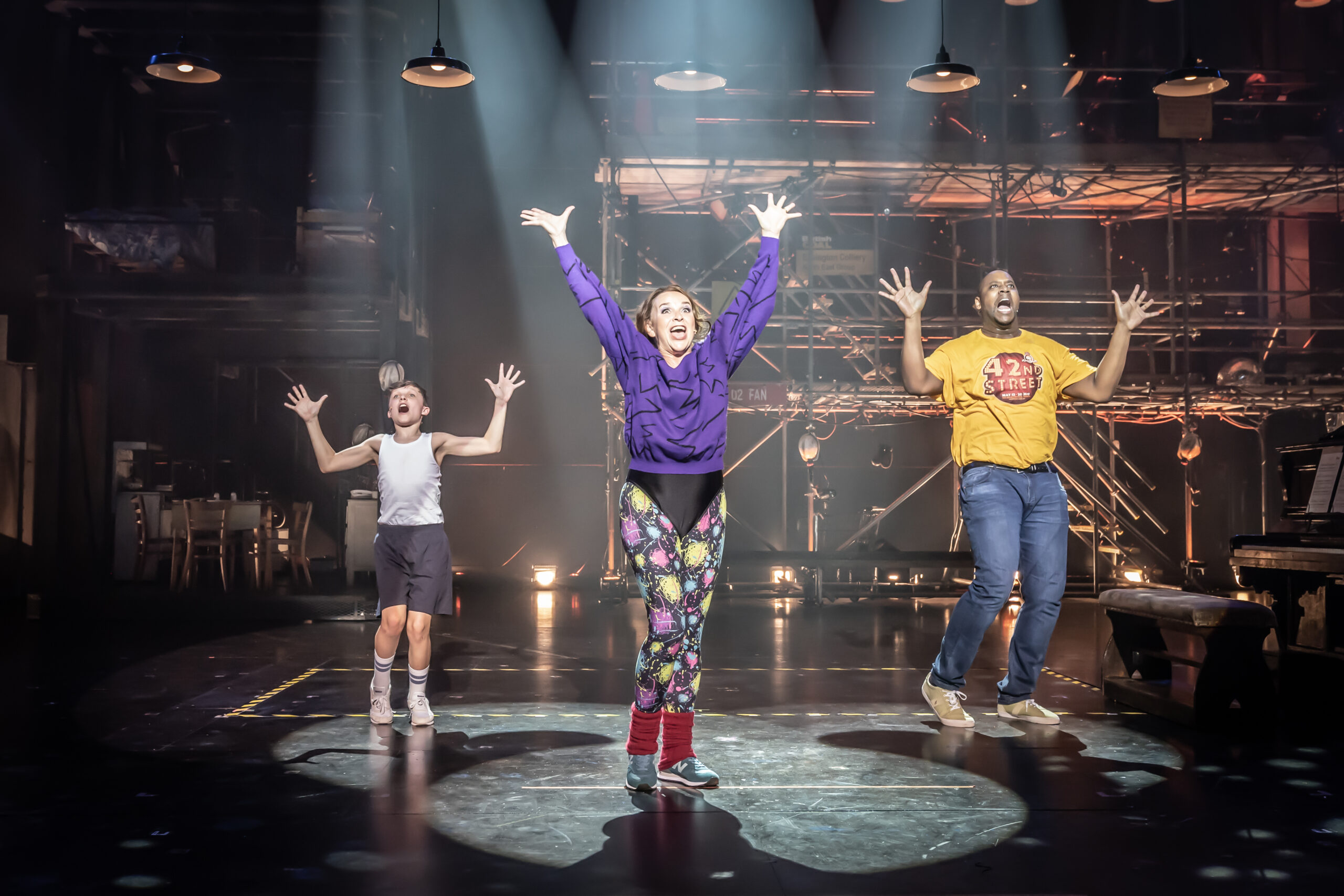 Production photograph from Billy Elliot. Billy Elliot (Jaden Shentall-Lee), Mrs Wilkinson (Sally Ann-Triplett) and Mr Braithwaite (Cameron Johnson) dance together jubilantly in the community hall. Billy and Mrs Wilkinson wear 1980s workout clothes, and Mr Braithwaite wears jeans and a Musical Theatre t-shirt.