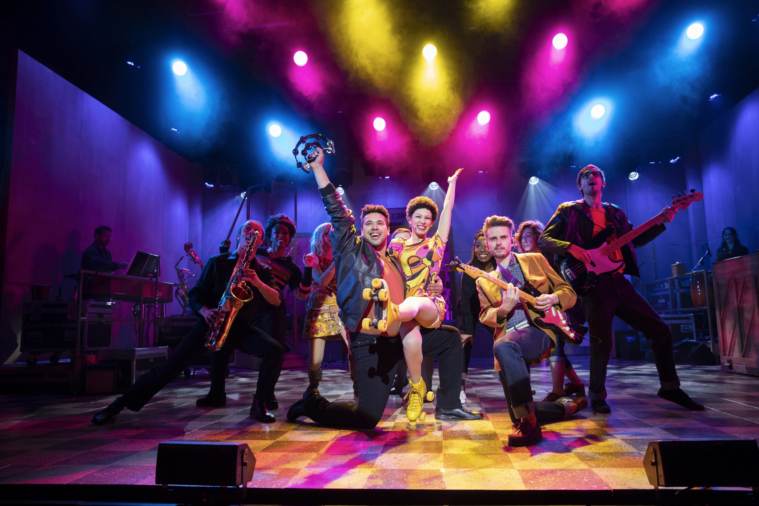 Production photograph from Beautiful - The Carole King Musical. Little Eva (Amena El-Kindy) smiles as she sits on the knee of a musician (Kevin Yates). The pair are smiling with an arm aloft, surrounded by other musicians and studio workers including Gerry Goffin (Tom Milner). Eva is wearing a 70s patterned dress and yellow roller skates, with cropped black hair, beneath candy-coloured lights in yellow, pink and blue.