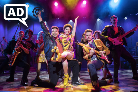 Production photograph from Beautiful - The Carole King Musical. Little Eva (Amena El-Kindy) smiles as she sits on the knee of a musician (Kevin Yates). The pair are smiling with an arm aloft, surrounded by other musicians and studio workers including Gerry Goffin (Tom Milner). Eva is wearing a 70s patterned dress and yellow roller skates, with cropped black hair, beneath candy-coloured lights in yellow, pink and blue. There is a white AD - Audio Description - logo top left.