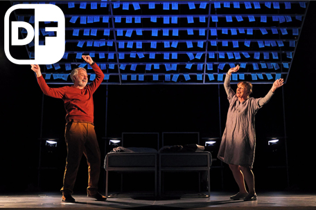 Production image from Maggie May. Maggie (Eithne Brown) and Gordon (Tony Timberlake) dance with glee around their bed beneath a ceiling covered in blue post-it notes, their top halves illuminated in warm spotlights. There is a white DF - Dementia Friendly - logo top left).