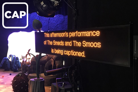 A caption unit pictured next to the empty stage for The Smeds and The Smoos, Yellow LED text on the black unit reads 'This afternoon's performance of The Smeds and The Smoos is being captioned.' There is a white CAP logo top left.