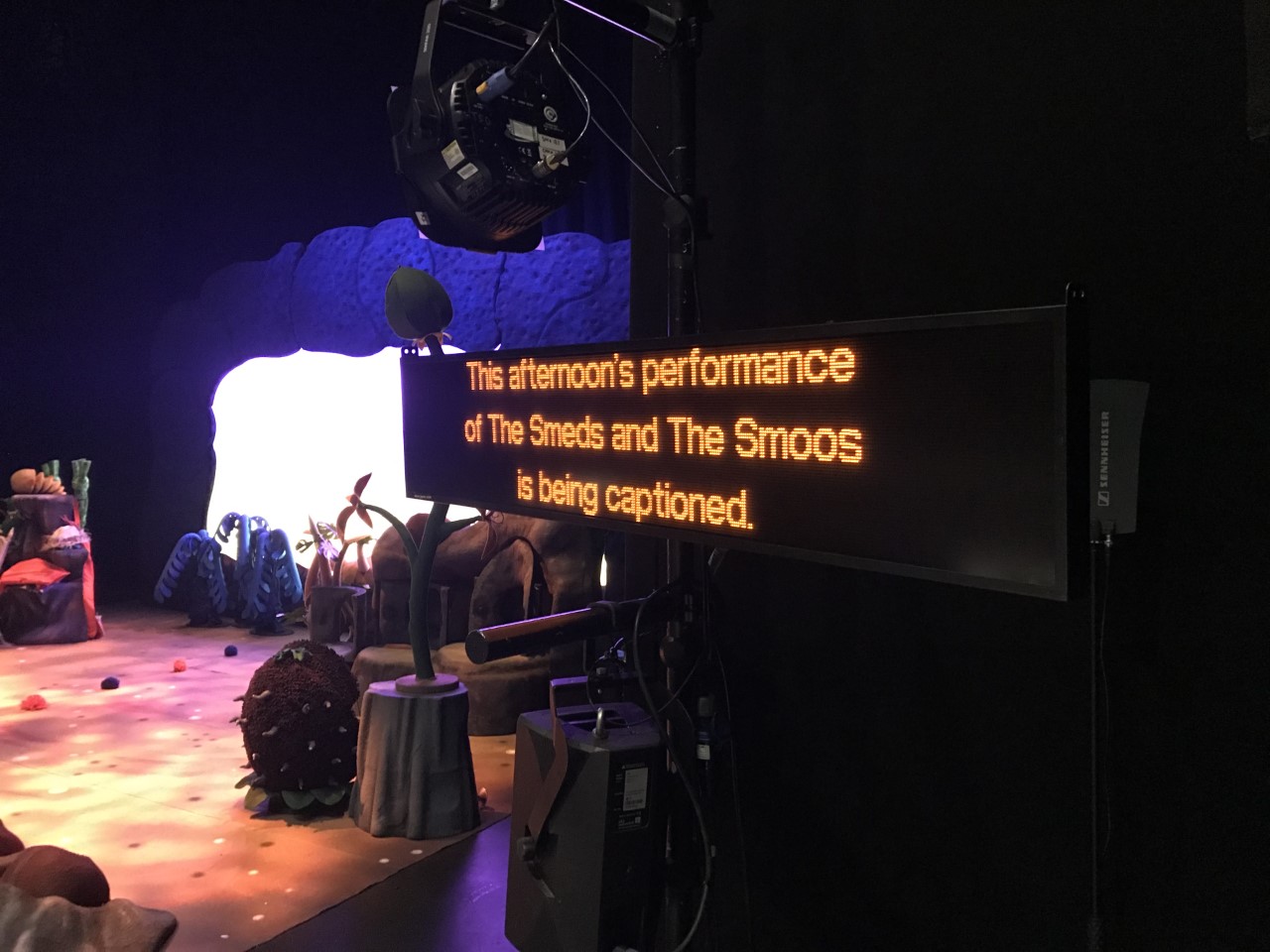 A caption unit pictured next to the empty stage for The Smeds and The Smoos, Yellow LED text on the black unit reads 'This afternoon's performance of The Smeds and The Smoos is being captioned.'