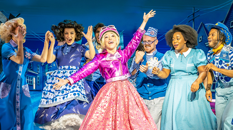 Production image from The Wizard of Oz. Glinda the Good Witch - wearing a shiny pink jacket, a sparkly pink a-line skirt and a pink helmet with a blonde ponytail - exclaims with her arms aloft. Four munchkins and Dorothy, all wearing blue toned outfits, watch on with joyful expressions.