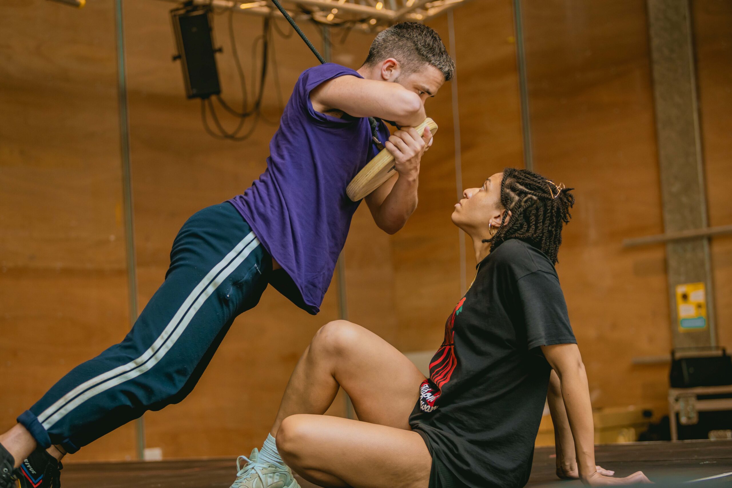 In a wood-cladded rehearsal space, Felipe Pacheco (Gregor) leans over Hannah Sinclair Robinson (Grete), supported by a swing. Hannah looks up at him from her seated position on the floor, her arms behind her for support. Both are wearing casual leisurewear.