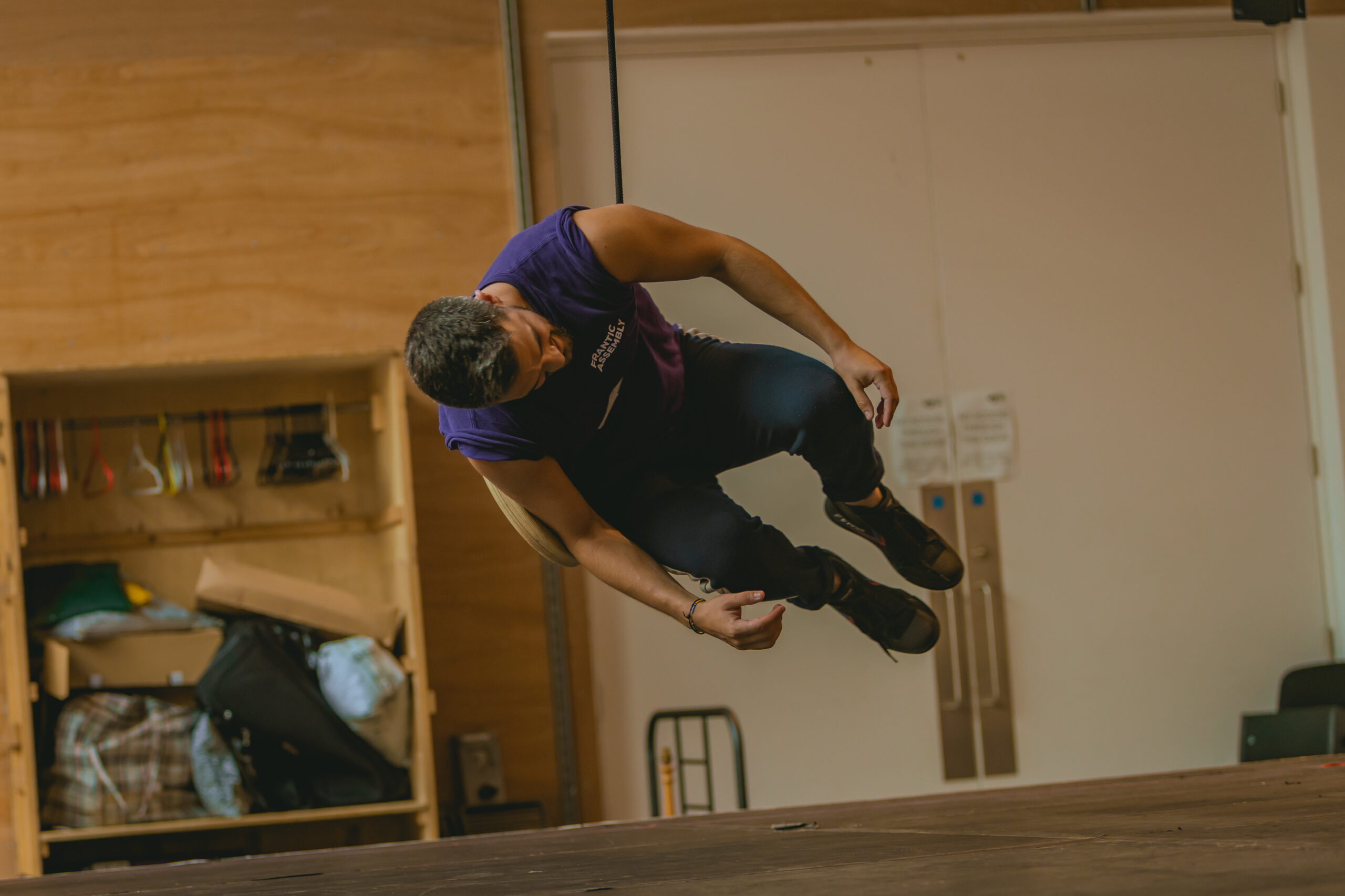 In a wood-cladded rehearsal room, Felipe Pacheco (Gregor) is swinging in the air facing the ground, with the rope swing wrapped around his torso.