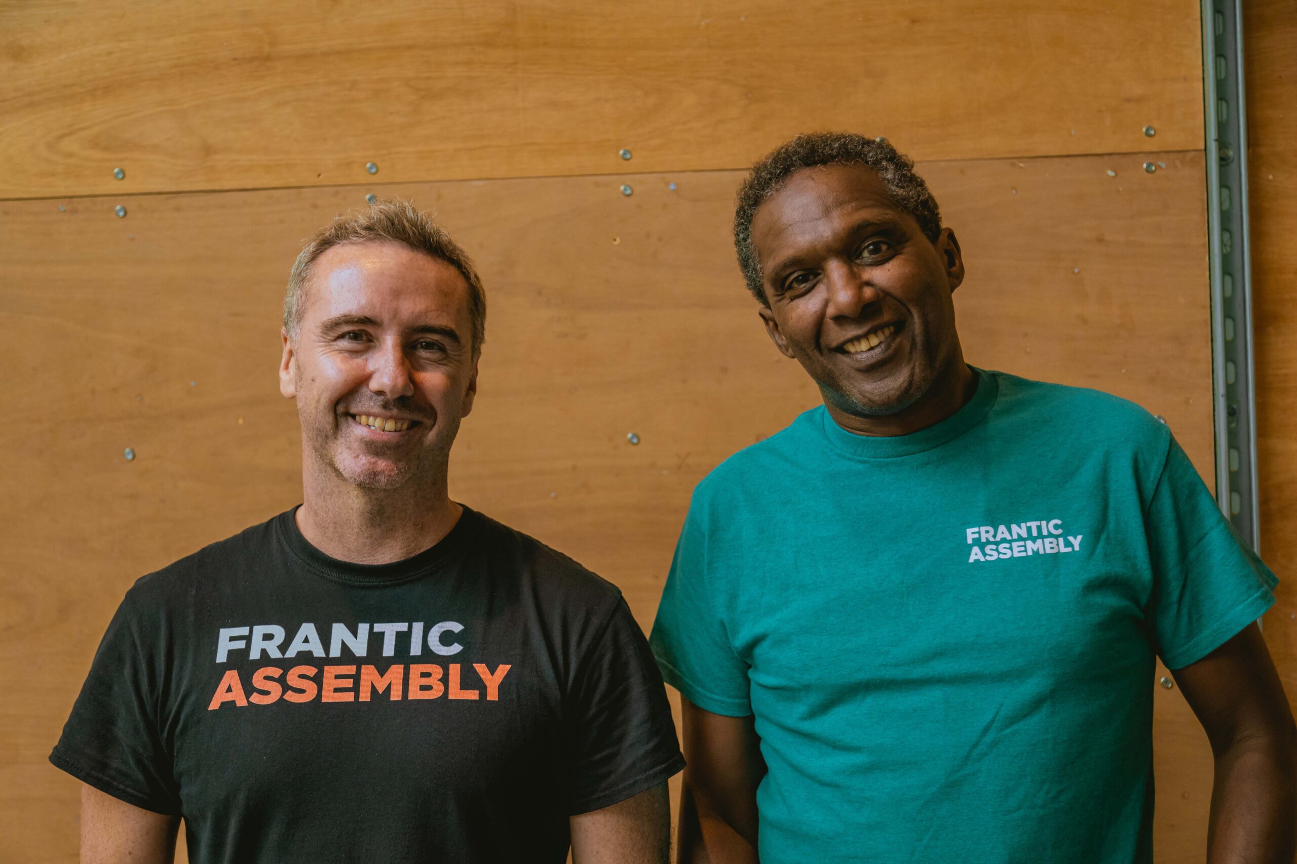 Frantic Assembly Artistic Director Scott Graham and Adaptor Lemn Sissay OBE smile against a wood-cladded wall. They are pictured from the torso up, and both are wearing t-shirts with Frantic Assembly branding. Scott's t-shirt is black, with a grey and orange logo across the chest, and Lemn's is teal, with a white logo on the left of the chest.