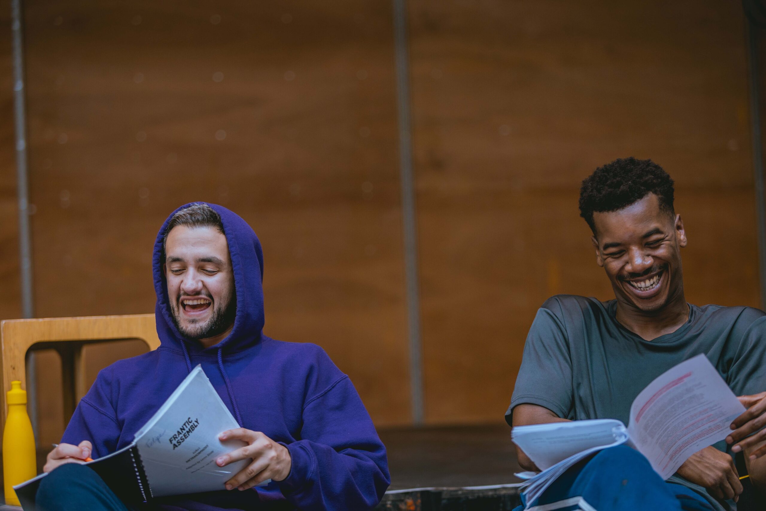 In a wood-cladded rehearsal room, Felipe Pacheco (Gregor) and Troy Glasgow (Father) are sitting together discussion the scripts in their hands. Both are laughing with happy expressions.