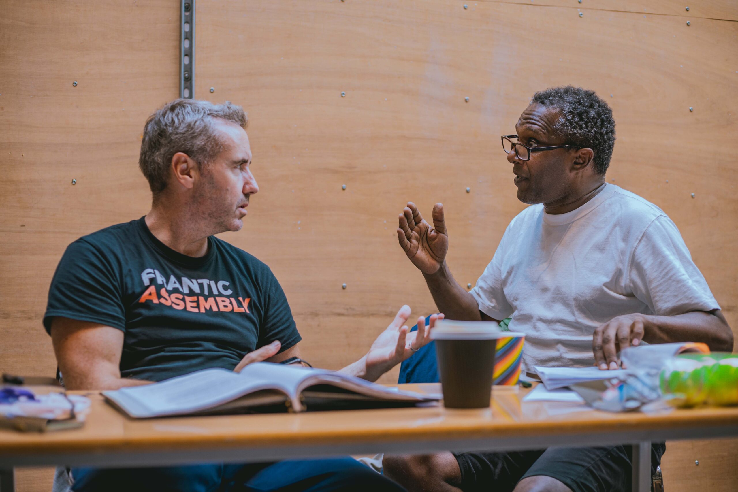 In a wood-cladded rehearsal room, Frantic Assembly Artistic Director Scott Graham is in discussion with Adaptor Lemn Sissay OBE. Both are deep in discussion with one another. Scott is wearing a black t-shirt with the Frantic Assembly logo while Lemn is wearing a plain white t-shirt.