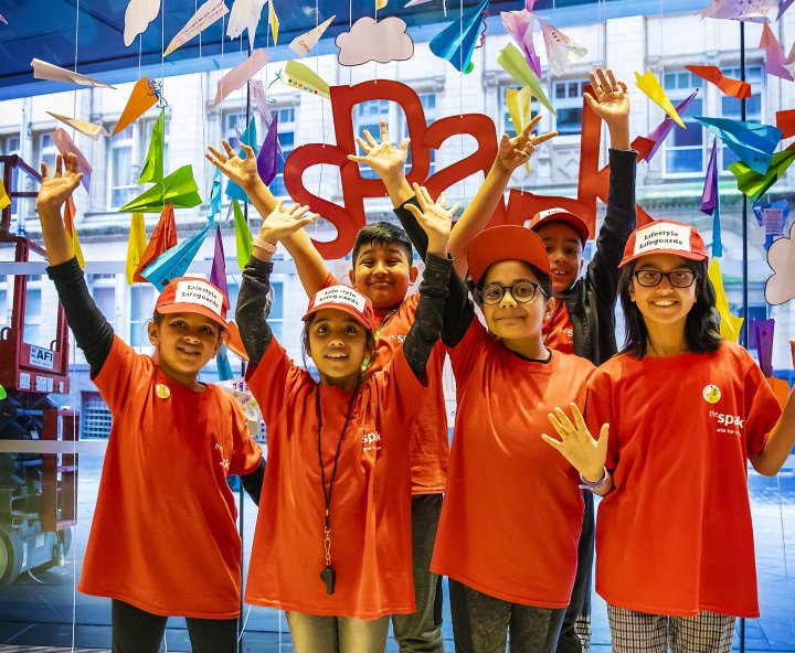 Six children stand together in a group at the Spark Takeover Day 2022. They are smiling with their arms aloft, wearing red t-shirts and caps in front of a backdrop of paper planes.