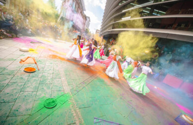 A group of South Asian dancers perform on Orton Square amongst plumes of colourful holi paint. The dancers are wearing long white dresses and colourful fabric sashes, as an adoring crowd watch on.