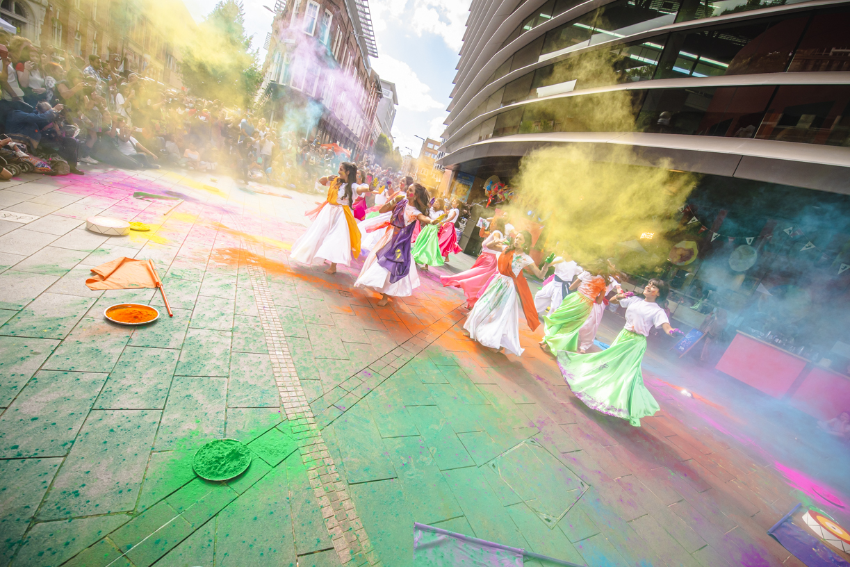A group of South Asian dancers perform on Orton Square amongst plumes of colourful holi paint. The dancers are wearing long white dresses and colourful fabric sashes, as an adoring crowd watch on.